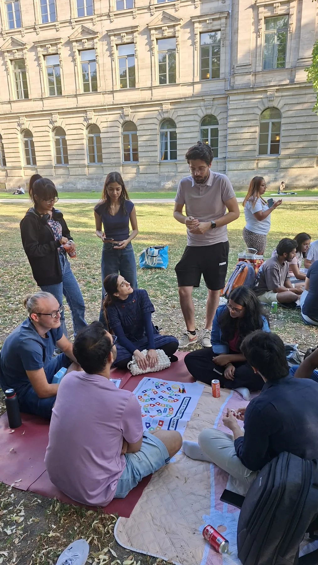 Guests sitting around the Invest in Youth board game in a park in Erlangen, with a historic building behind them.
