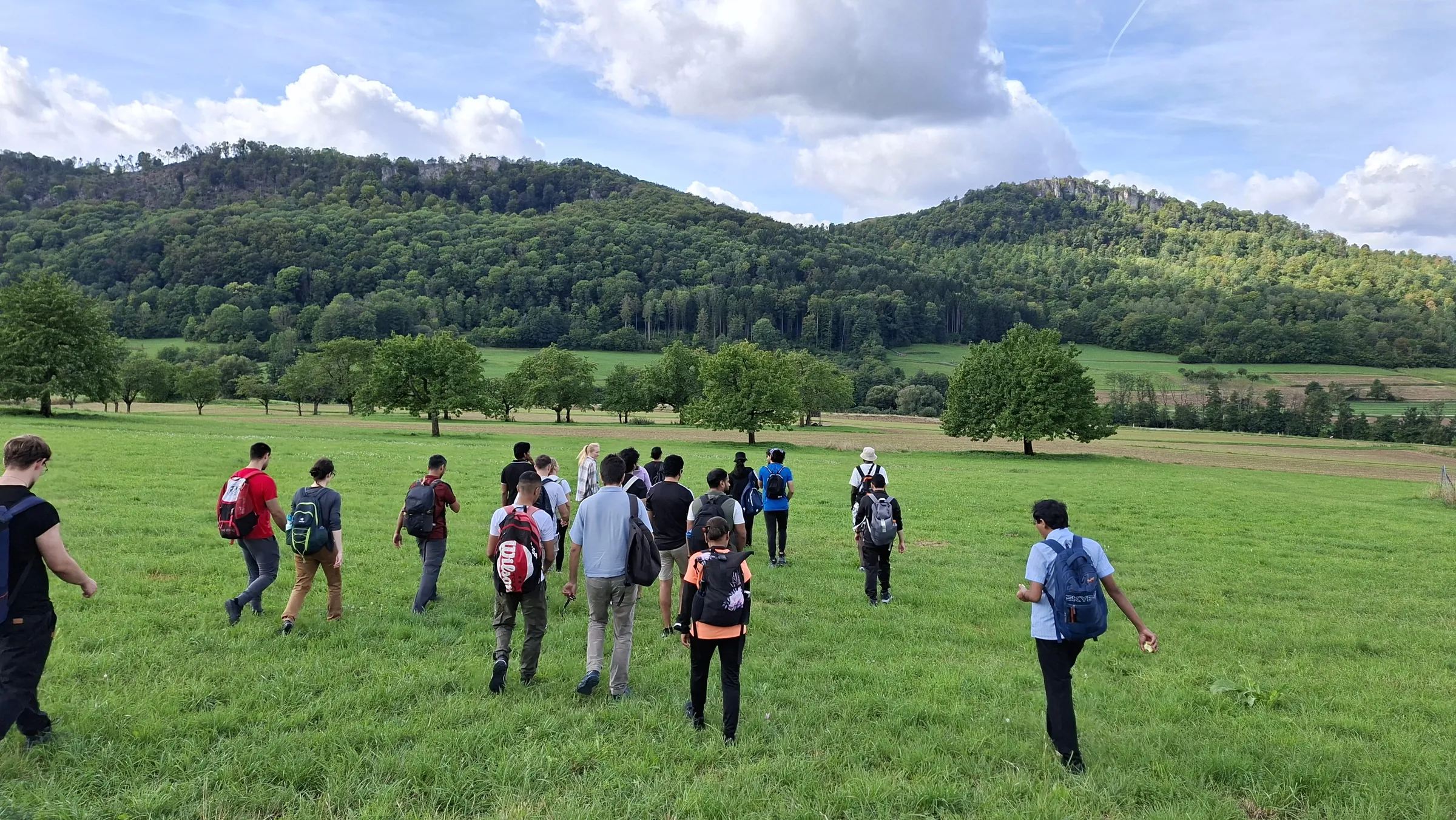 A group of people with backpacks walks across a wide green meadow toward a line of trees and forested hills.