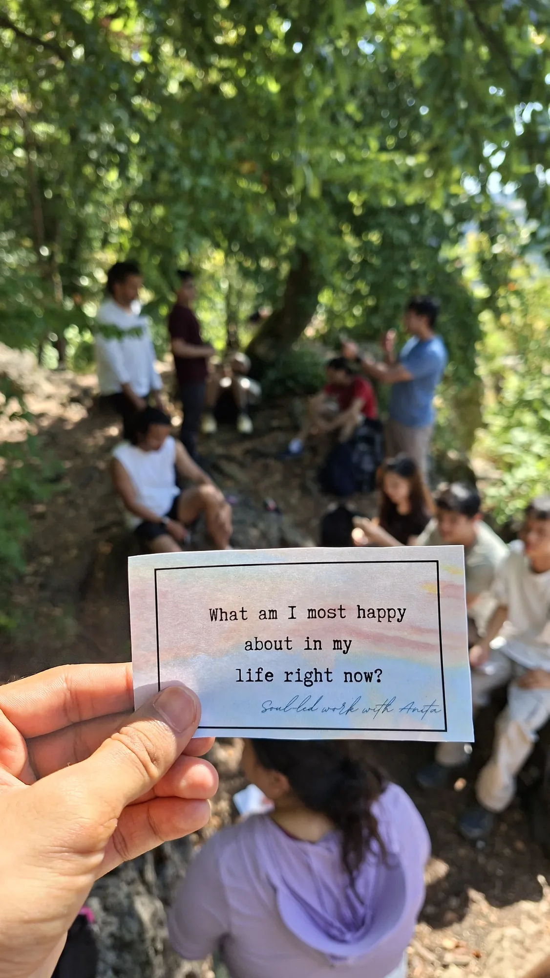 A hand holds a card asking what the holder is most happy about in life right now, while a group sits under trees behind it.