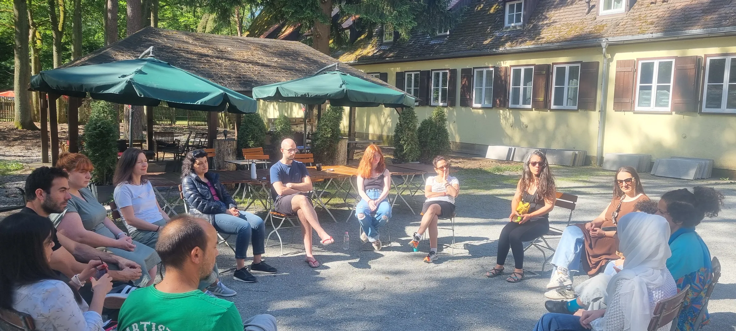 People sit in a loose circle of chairs in a sunny courtyard beside a pale yellow building and green umbrellas.